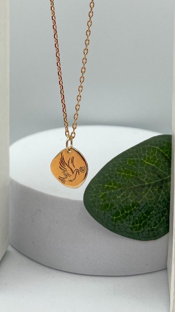 Collier en plaqué or avec pendentif colombe gravée et rameau d’olivier, posé sur support blanc et feuille verte