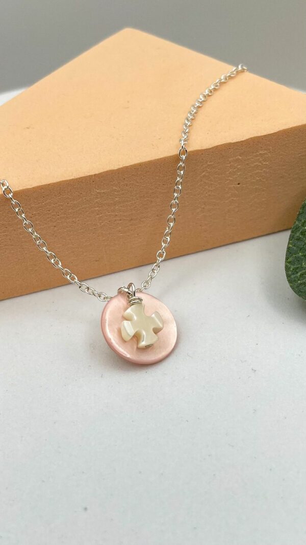 Collier catho pour première communion avec croix en nacre blanche et pastille rose sur chaîne en argent, posé sur fond beige