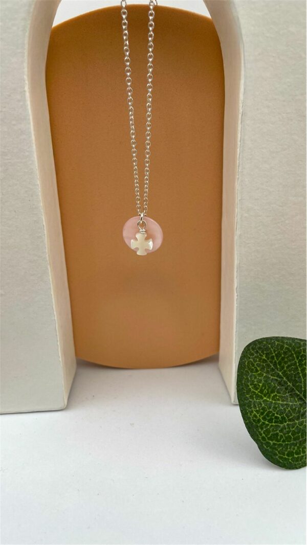 Collier religieux pour jeune fille suspendu verticalement, croix blanche en nacre sur disque rose, fond beige et blanc